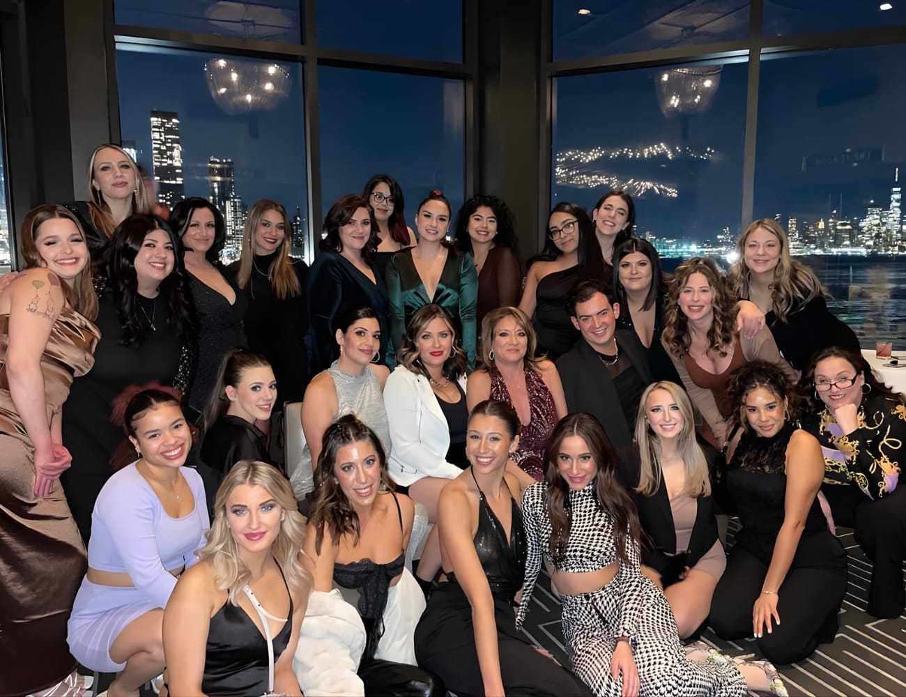 Group of people posing indoors with a city skyline and fireworks visible in the background.