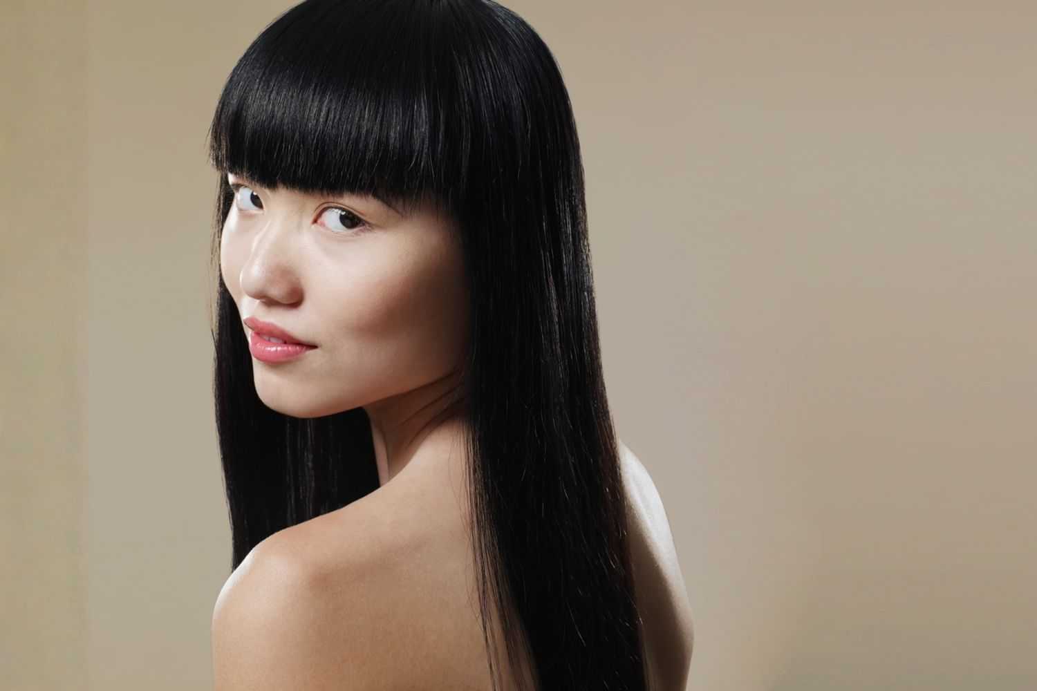 A woman with long, black hair and bangs looks over her shoulder against a beige background.