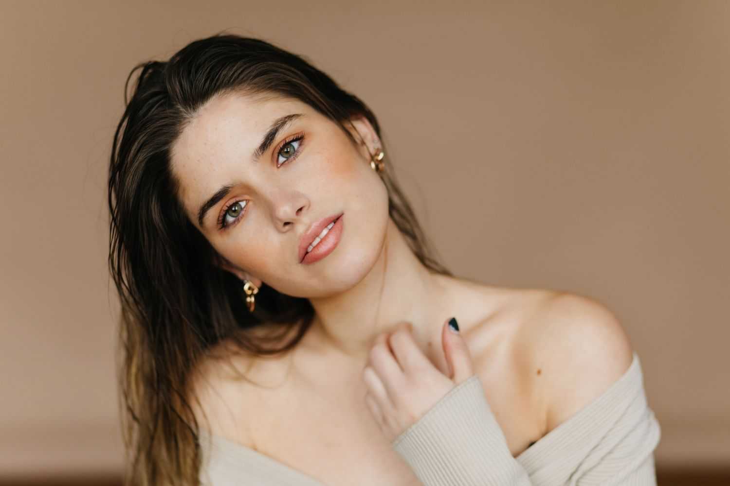 Young woman gazing softly, wearing an off-shoulder top with natural makeup and earrings.